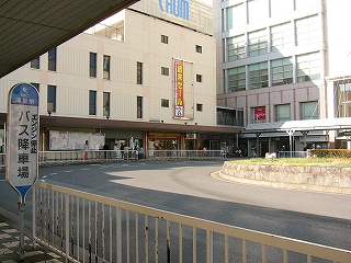 奥に津駅駅舎の端の部分、右にアスト津の建物の側面。それらに囲まれるところへ右カーブを描いて入り込むロータリーの一部。駅舎前では多くの人がバスを待っている。