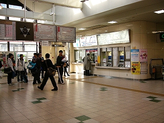 壁に埋め込まれるように設置された券売機の並びを右斜めに見て。床面は白色の大きい正方形の化粧板が敷き詰められていて、そこに時折緑色の化粧板が混じる。上方には電照式時刻表と時計が吊られている。