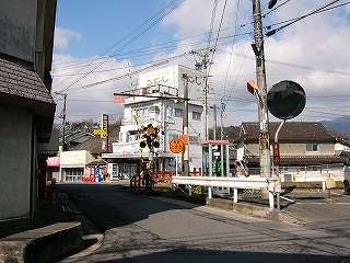 1.5車線ほどの細い道路の先に踏切、そして4階建ての高さの白い建物一件。
