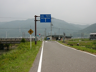 細い道路。上に青い道案内の看板。左は川、右は畑。
