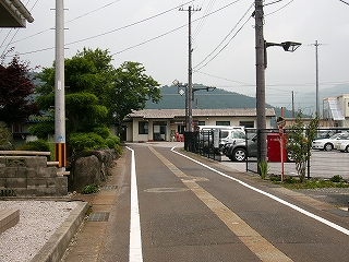 左に背の高い緑のある人家、右手はフェンスに囲われた駐車場、少し遠くの突き当たりに平屋の駅舎。