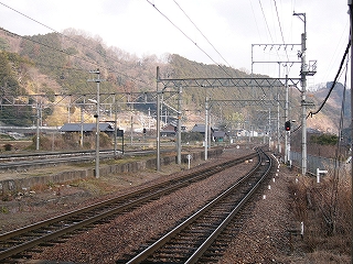 遠くに山。トラス架線柱の跨ぐ線路が伸びる。