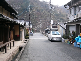 1.5車線ほどの道路。両脇に民家。遠く突き当たりは山肌でＴ字路になっている。
