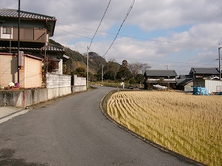 自動車一台分ぐらいの道路。左手に薄茶色の切り株だけになった田んぼ。左手に民家。